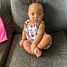 baby, child, sitting, couch, gray_couch, hair_clips, red_clips, colorful_outfit, ice_cream_pattern, cross_legged, curious_expression, indoors, furniture, soft_light, portrait, person, young_child, cute, skin, face