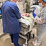 newborn, baby, hospital, medical_staff, scrubs, mask, gloves, medical_equipment, crib, infant, healthcare, clinic, delivery_room, person, face, protective_gear, blue_shoes, white_floor, towel, hospital_gown