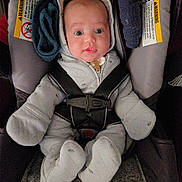 Toby is registered to the contest to win money with this photo: baby, infant, car_seat, onesie, hooded_clothing, straps, safety, seat, child, young, cute, indoor, person, face, wide_eyes, clothing, seatbelt, warning_label, soft, expression