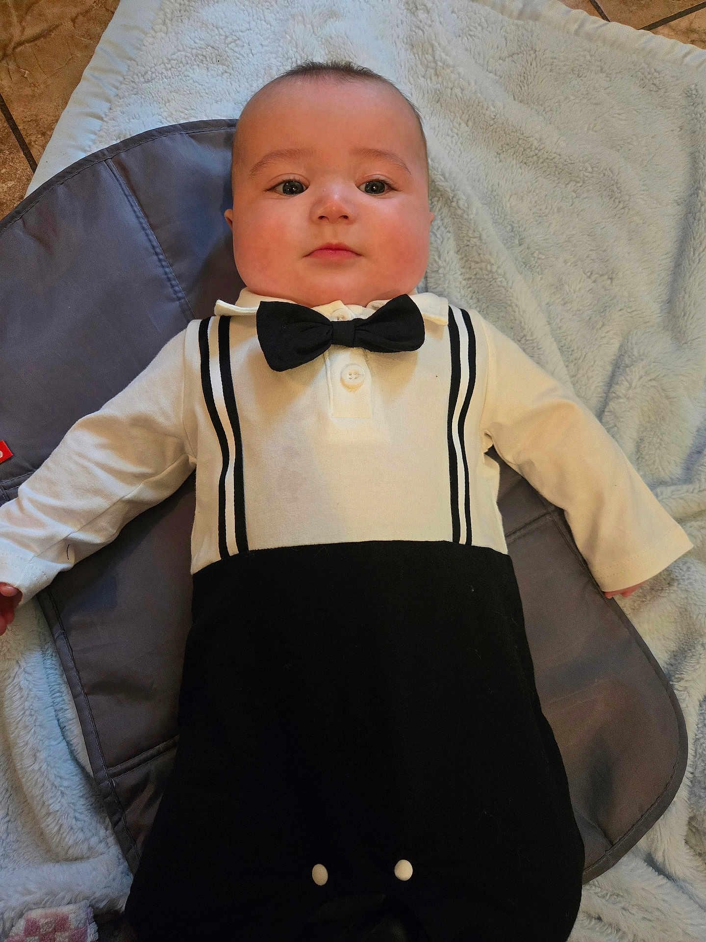 Toby joined the competition — help win amazing prizes! baby, infant, child, bow_tie, onesie, blanket, clothing, face, chubby_cheeks, lying_down, portrait, eyes, hand, outfit, tile_floor, soft_texture, fabric, neutral_expression, gaze, indoor