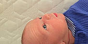 Toby is registered to the contest to win money with this photo: baby, newborn, infant, face, head, blue_clothing, ribbed_fabric, striped_collar, skin, ear, eye, nose, mouth, lying_down, textured_surface, mattress, portrait, close_up, child, curious