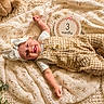 baby, infant, smiling, tongue_out, headband, overalls, blanket, teddy_bear, greenery, laying_down, cute, child, three_months, indoors, cozy, happy, portrait, soft_light, fabric, decor