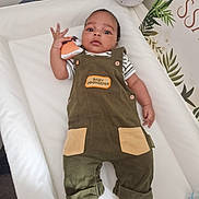 Khalil is registered to the contest to win money with this photo: baby, infant, child, overalls, toy, nursery, cushion, white, green, clothing, person, footwear, barefoot, face, looking, lying_down, cute, indoor, expression, decor