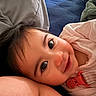 baby, infant, child, portrait, close_up, wide_eyes, lying_down, smiling, face, dark_hair, pink_clothing, romper, red_buckle, blanket, bed, pillow, plush_toy, indoor, cozy, photography
