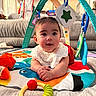 baby, child, infant, tummy_time, smiling, sitting, playmat, play_gym, toys, colorful, indoors, living_room, couch, portrait, face, hands, bib, baby_clothing, soft_toys, happy