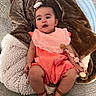 baby, child, infant, sitting, couch, blanket, pillow, portrait, smiling, open_mouth, flower_hair_clip, pink_outfit, bib, dress, sandals, sunflower, pacifier, indoors, home, happy