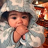 animal_ears, baby, blue_clothing, child, close_up, cozy, cute, dining_room, fuzzy_hood, hand_in_mouth, hooded_outfit, indoors, infant, portrait, restaurant, sitting, warm_lighting, white_clothing, wide_eyes, wooden_chairs