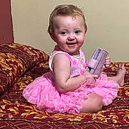 Oaklynn is registered to the contest to win money with this photo: baby, child, pink_dress, smiling, sitting, bed, purse, indoor, cute, infant, face, happy, toddler, patterned_bedspread, furniture, person, young_child, playful, holding_object, portrait