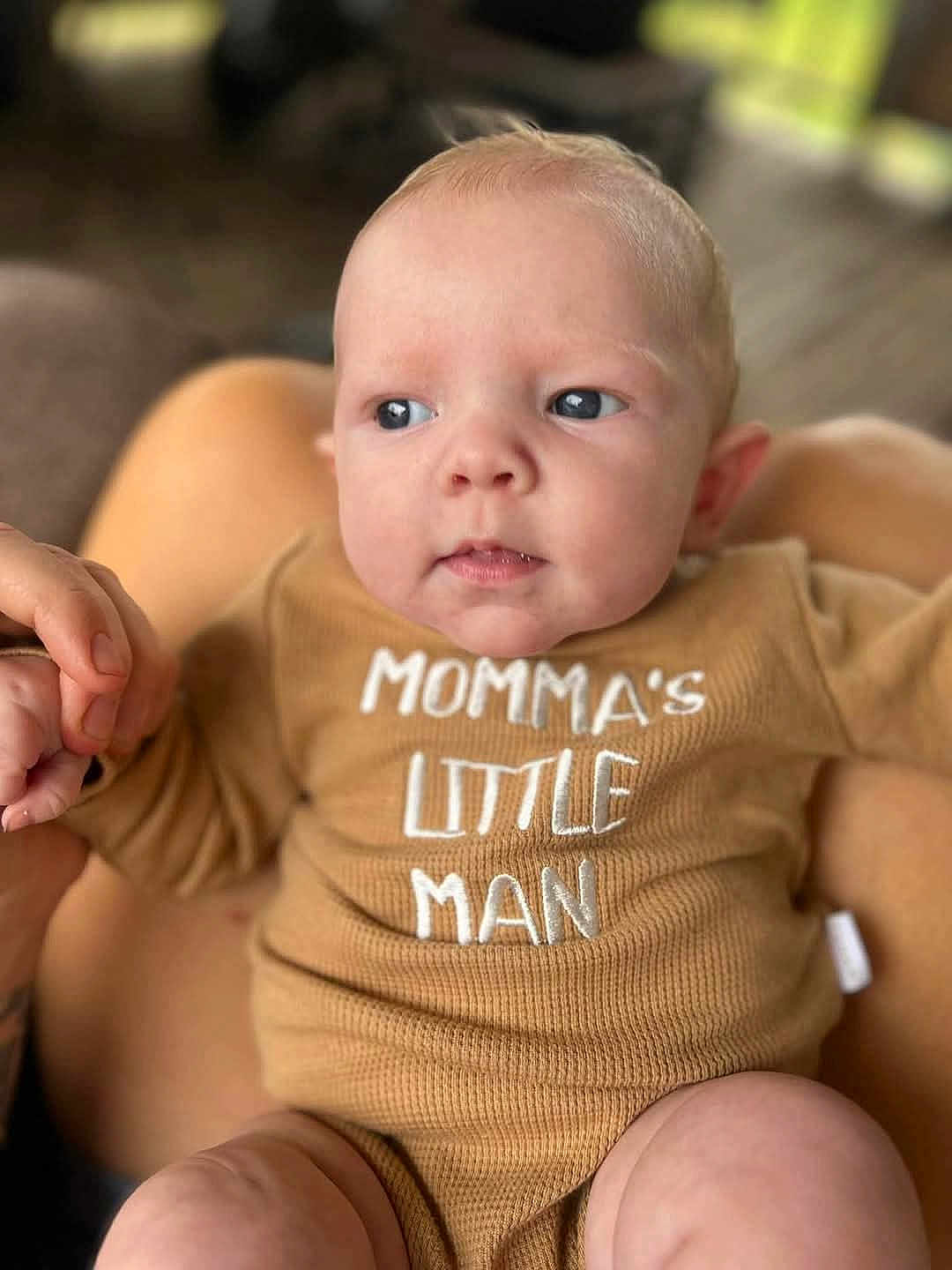 Kolton joined the competition — help win amazing prizes! baby, infant, onesie, clothing, hand, holding, face, blue_eyes, skin, person, closeup, expression, small_fingers, blurred_background, indoor, cute, child, newborn, human, portrait