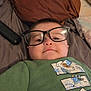 Antonio is registered to the contest to win money with this photo: child, toddler, glasses, eyeglasses, bed, pillow, blanket, green_shirt, graphic_print, smile, face, portrait, indoor, remote_control, bedding, cozy, cute, lying_down, toddler_clothing, short_hair