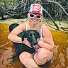 woman, dog, water, sunglasses, cap, smile, outdoor, nature, tree_roots, riverbank, summer, pet, black_dog, happy, sitting, swimsuit, sunny, playful, friendship, relaxation