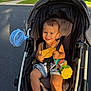 toddler, stroller, leaf, smiling, child, outdoor, sunlight, fan, toy, shorts, seatbelt, happy, summer, greenery, residential, sidewalk, asphalt, playful, baby, parked_vehicle