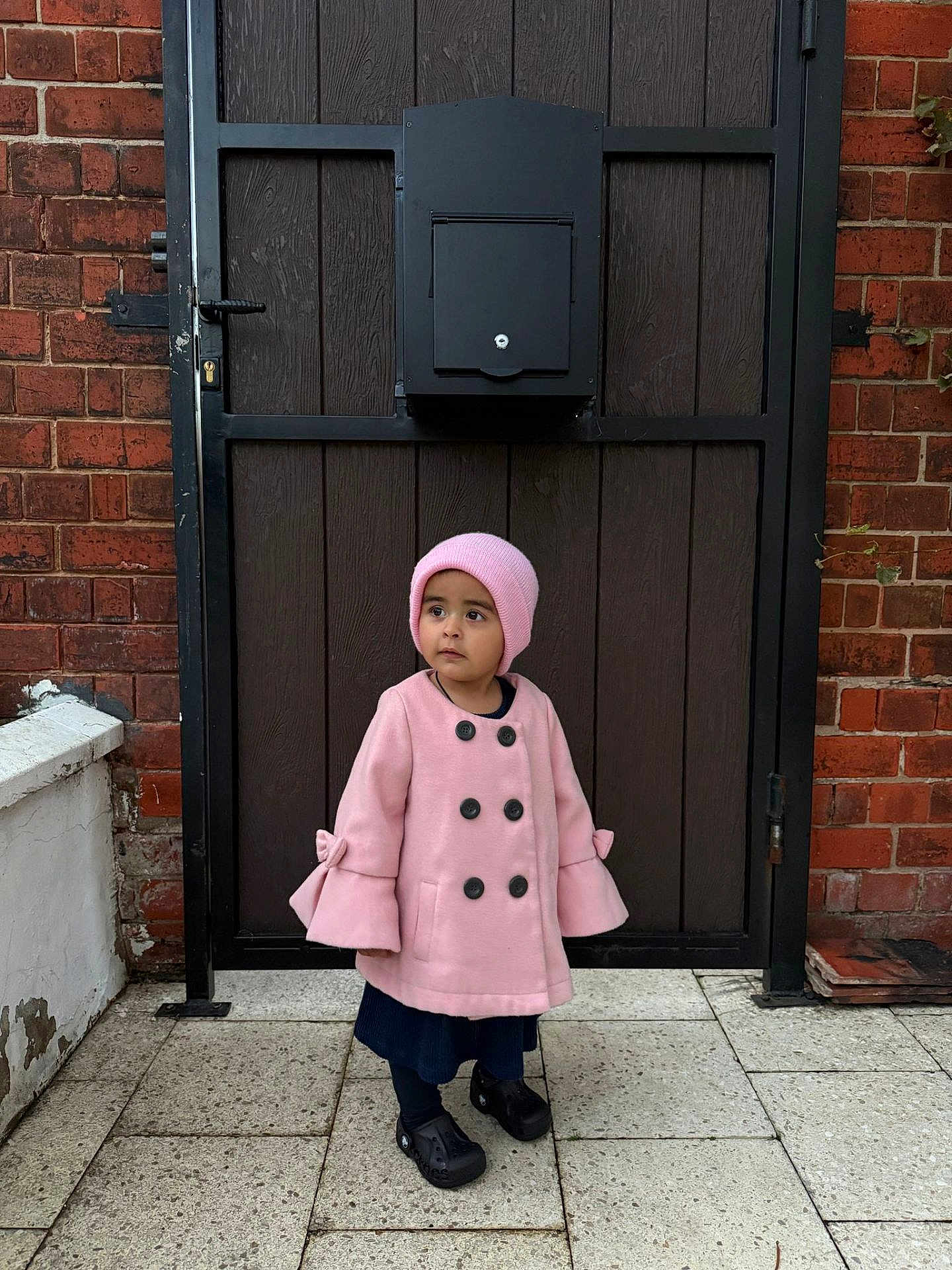 Inayah is registered to the contest to win money with this photo: child, pink_coat, pink_hat, black_shoes, brick_wall, wooden_door, mailbox, outdoor, pavement, side_view, standing, curious, winter_clothing, young_child, portrait, daylight, urban, casual, fashion, person