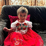 Dua Zara joined the competition — help win amazing prizes! child, toddler, smiling, red_dress, white_top, embroidery, sitting, couch, leather_couch, cushion, curtains, indoors, natural_light, happy, person, face, hair, fabric, pattern, furniture