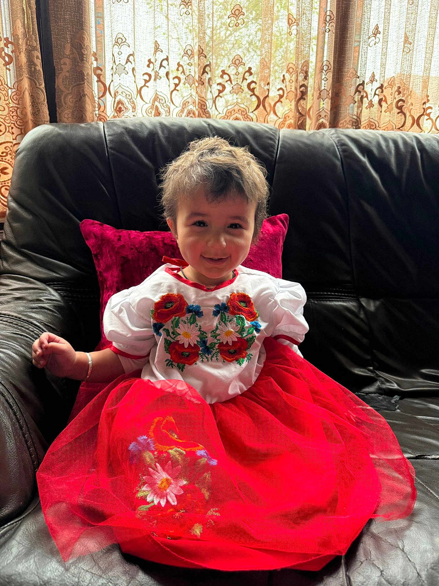 Dua Zara joined the competition — help win amazing prizes! child, toddler, smiling, red_dress, white_top, embroidery, sitting, couch, leather_couch, cushion, curtains, indoors, natural_light, happy, person, face, hair, fabric, pattern, furniture
