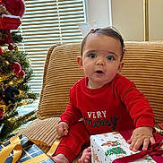 Amira joined the competition — help win amazing prizes! baby, child, christmas_tree, present, gift_wrap, ribbon, red_pajamas, couch, holiday, holiday_decor, indoor, festive, curious, smiling, face, person, sitting, window_blinds, home, celebration
