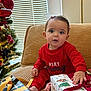baby, child, christmas_tree, present, gift_wrap, ribbon, red_pajamas, couch, holiday, holiday_decor, indoor, festive, curious, smiling, face, person, sitting, window_blinds, home, celebration