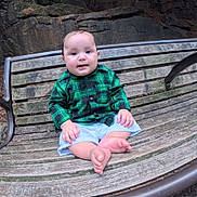 Wyatt is registered to the contest to win money with this photo: baby, child, infant, sitting, bench, outdoor, park, forest, rocks, plaid_shirt, green_clothing, white_shorts, barefoot, smiling, portrait, face, hands, legs, wooden_bench, nature