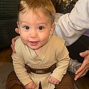 Anakin is registered to the contest to win money with this photo: adult_support, baby, beige, boots, bright_eyes, brown, child, clothing, costume, cute, floor, hands, happy, indoors, infant, person, portrait, sitting, smile, young_child