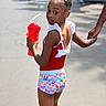 beads, casual_clothing, child, colorful_shorts, daytime, expression, girl, hair_bun, hand_holding, outdoor, people, red_cup, red_top, side_view, straw, street, summer, sunlight, urban, walking
