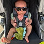 baby, stroller, sunglasses, toy, dinosaur, infant, child, seatbelt, cute, outdoor, summer, shade, foot, hand, pink_clothing, green_toy, black_stroller, sunlight, reflection, casual
