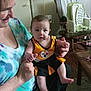 baby, woman, indoor, smiling, holding, sportswear, pittsburgh_steelers, rocking_chair, lamp, table, carpet, wood_floor, furniture, person, child, casual_clothing, home, living_room, portrait, seated