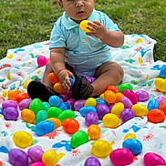 Ryland joined the competition — help win amazing prizes! baby, child, grass, blanket, colorful, easter_eggs, plastic_eggs, outdoor, playing, sitting, dinosaur_pattern, footwear, sandals, blue_shirt, curious, cute, person, nature, spring, holiday
