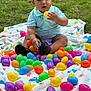 baby, child, grass, blanket, colorful, easter_eggs, plastic_eggs, outdoor, playing, sitting, dinosaur_pattern, footwear, sandals, blue_shirt, curious, cute, person, nature, spring, holiday