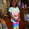 child, smiling, indoors, colorful_clothing, donut_graphic, rainbow_pants, standing, wooden_floor, furniture, living_room, cabinet, sneakers, hair_bow, happy, person, portrait, home, casual, young_girl, cute