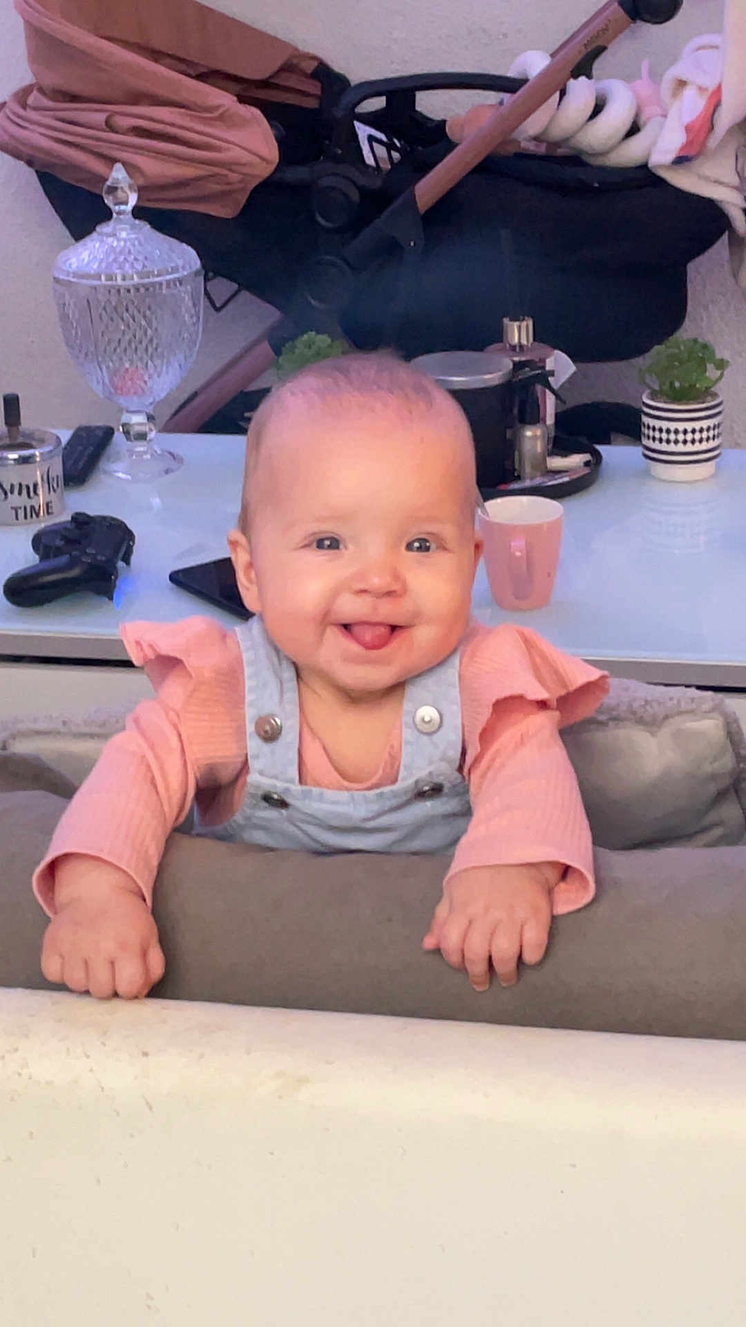 Mya a rejoint le concours — aidez-le/la à gagner de superbes lots ! baby, child, smiling, tongue_out, indoor, cushion, pink_clothing, overalls, stroller, table, cup, gaming_controller, plant, decor, face, person, happy, infant, young_child, furniture
