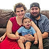 family, baby, woman, man, couch, smile, happy, indoor, casual_clothing, red_dress, cap, beard, parent, child, portrait, seated, leather_furniture, tattoo, short_hair, holding