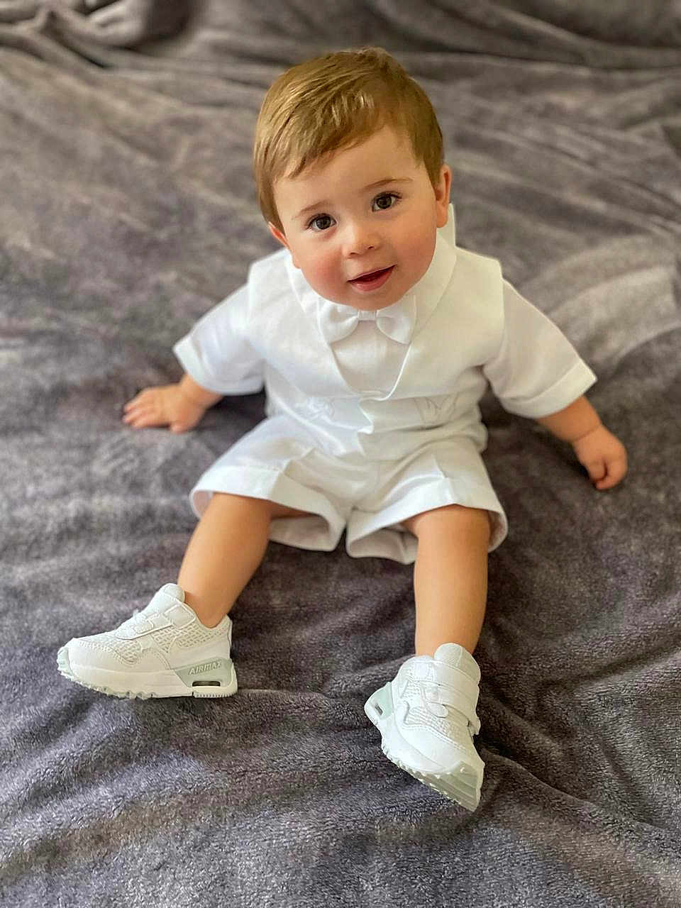 Lucca is registered to the contest to win money with this photo: toddler, child, white_outfit, bow_tie, sneakers, sitting, gray_blanket, cute, portrait, indoors, baby, smiling, happy, young_child, fashion, footwear, soft_texture, casual, person, closeup