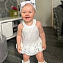 baby, bow, carpet, child, cute, fashion, furniture, happy, headband, home_interior, indoor, living_room, person, portrait, smiling, socks, standing, toddler, white_dress, wooden_floor