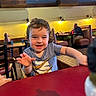 Hudson joined the competition — help win amazing prizes! child, toddler, smiling, curly_hair, grey_shirt, restaurant, table, chair, indoor, dining, person, light, yellow_wall, window, booth, adult, food, cup, happy, playful