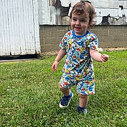 Hudson is registered to the contest to win money with this photo: toddler, child, grass, flower, smile, pajamas, dinosaur_print, sneakers, curly_hair, outdoor, building, weathered_wall, peeling_paint, greenery, playful, person, young_child, nature, summer, fun