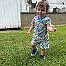 toddler, child, grass, flower, smile, pajamas, dinosaur_print, sneakers, curly_hair, outdoor, building, weathered_wall, peeling_paint, greenery, playful, person, young_child, nature, summer, fun