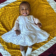 Kassia a rejoint le concours — aidez-le/la à gagner de superbes lots ! baby, blanket, child, cute, hands, happy, head, indoors, infant, lying_down, person, playful, portrait, skin, smiling, socks, textured_blanket, tongue_out, white_dress, yellow_blanket