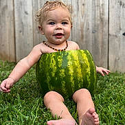 Oliver is registered to the contest to win money with this photo: baby, child, watermelon, grass, outdoor, summer, cute, smile, curly_hair, necklace, fence, fruit, green, nature, happy, sitting, playful, skin, barefoot, portrait