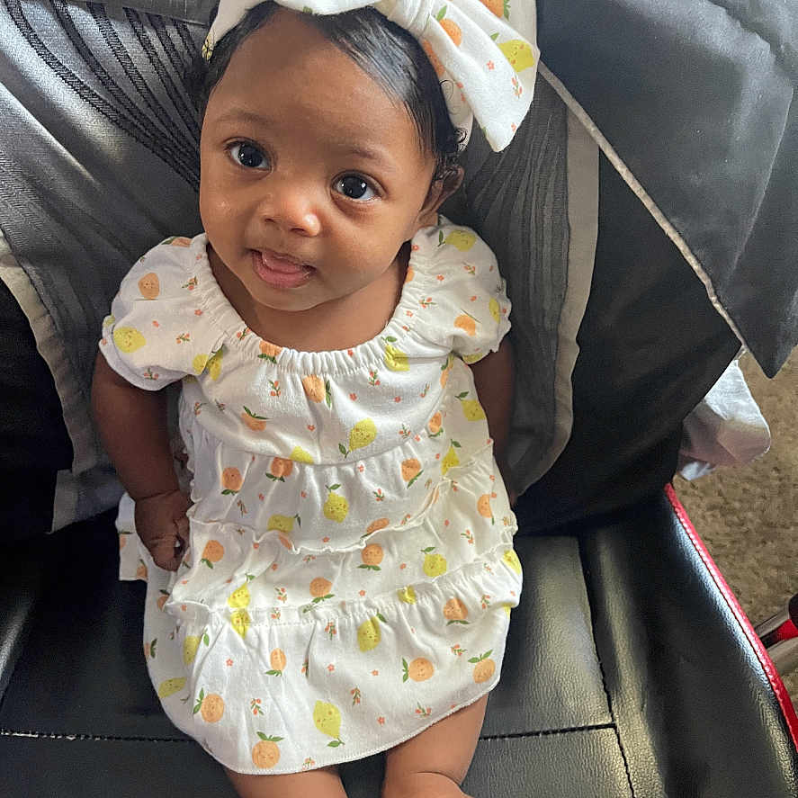 Azari joined the competition — help win amazing prizes! apparel, baby, black_leather_chair, bow, child, clothing, comfortable, curly_hair, cute, dress, fruit_pattern, happy, headband, indoors, infant, portrait, sitting, smiling, tongue_out, young_child