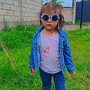 Alexandra is registered to the contest to win money with this photo: child, sunglasses, denim_jacket, jeans, boots, grass, outdoor, brick_wall, sky, person, serious, cute, fashion, casual, greenery, nature, standing, portrait, young, daytime