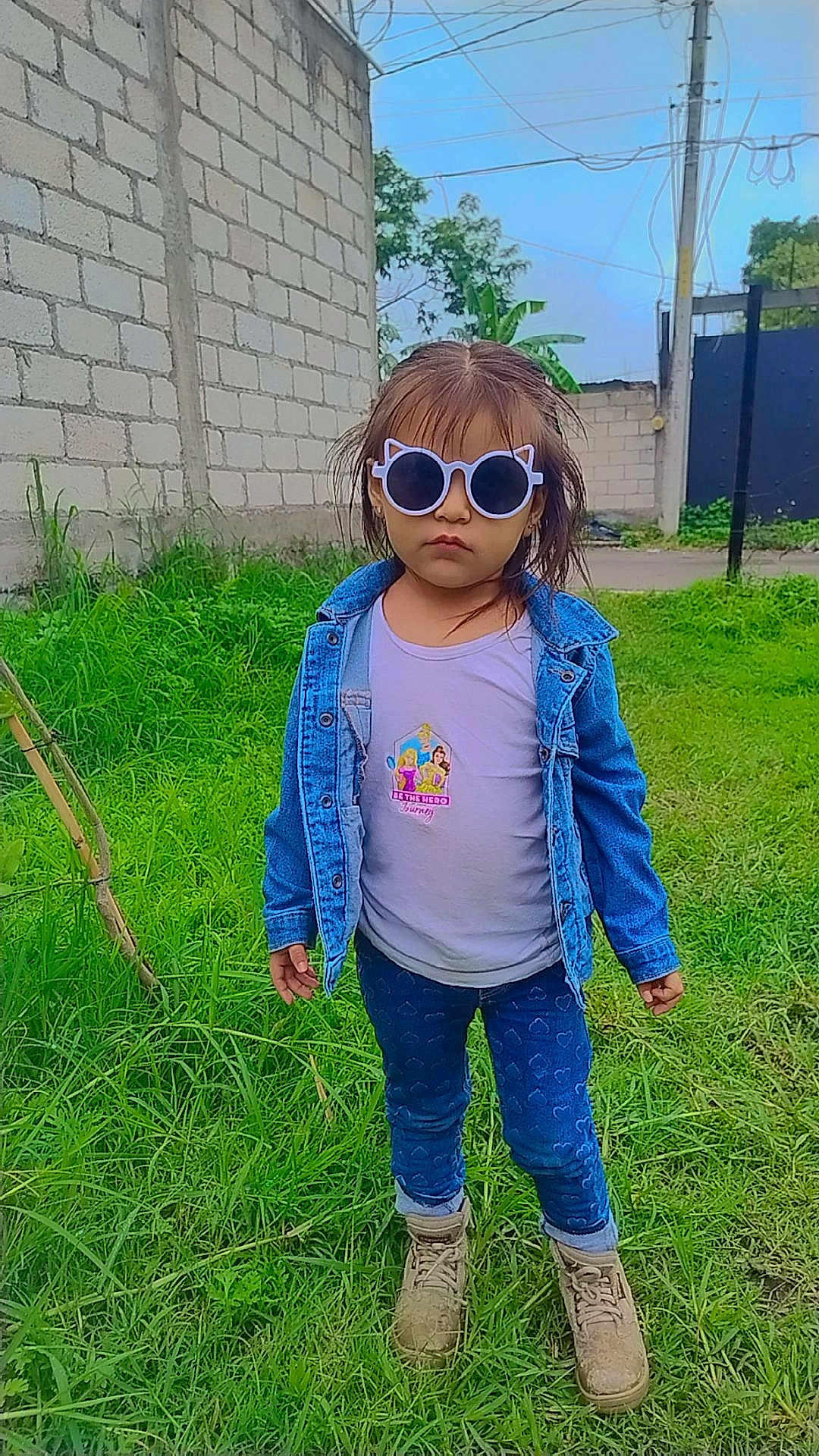 Alexandra is registered to the contest to win money with this photo: child, sunglasses, denim_jacket, jeans, boots, grass, outdoor, brick_wall, sky, person, serious, cute, fashion, casual, greenery, nature, standing, portrait, young, daytime