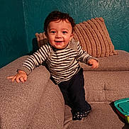 Aaron participe au concours pour gagner de l'argent avec cette photo : toddler, child, smiling, striped_shirt, curly_hair, couch, pillows, indoor, toy, happy, person, home, furniture, socks, jeans, texture, wall, living_room, playful, young_child