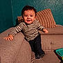 toddler, child, smiling, striped_shirt, curly_hair, couch, pillows, indoor, toy, happy, person, home, furniture, socks, jeans, texture, wall, living_room, playful, young_child