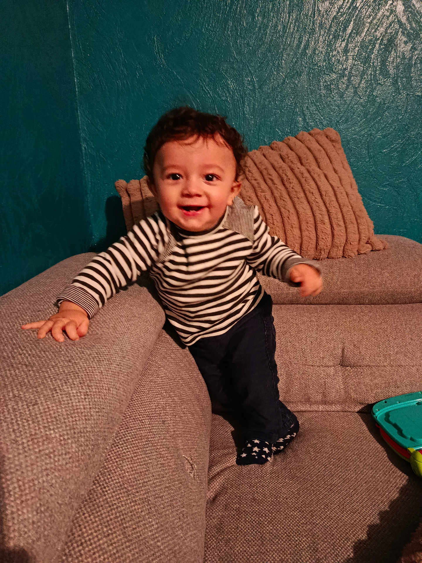 Aaron participe au concours pour gagner de l'argent avec cette photo : toddler, child, smiling, striped_shirt, curly_hair, couch, pillows, indoor, toy, happy, person, home, furniture, socks, jeans, texture, wall, living_room, playful, young_child