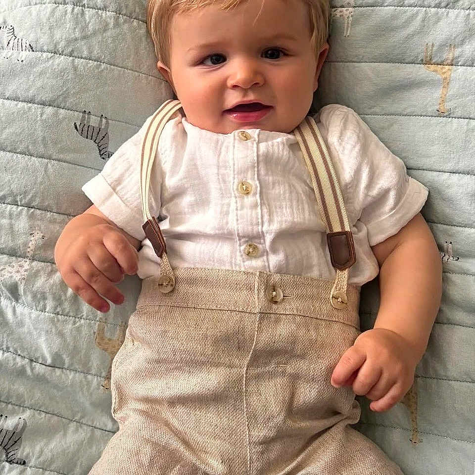 George joined the competition — help win amazing prizes! baby, child, smiling, face, clothing, suspenders, shorts, shirt, lying_down, quilt, blanket, cute, happy, infant, portrait, hand, light_brown_hair, indoors, soft_texture, fabric