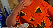 Christopher is registered to the contest to win money with this photo: child, orange_shirt, pumpkin_face, eating, table, plate, fork, indoor, dining_room, plant, picture_frame, bottle, wooden_chair, person, casual, food, seated, home, expression, young