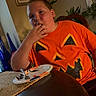 child, orange_shirt, pumpkin_face, eating, table, plate, fork, indoor, dining_room, plant, picture_frame, bottle, wooden_chair, person, casual, food, seated, home, expression, young