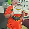 child, student, certificate, orange_shirt, jack_o_lantern, balloons, classroom, chair, decorations, award, proud, person, face, portrait, celebration, school, tshirt, indoor, standing, young