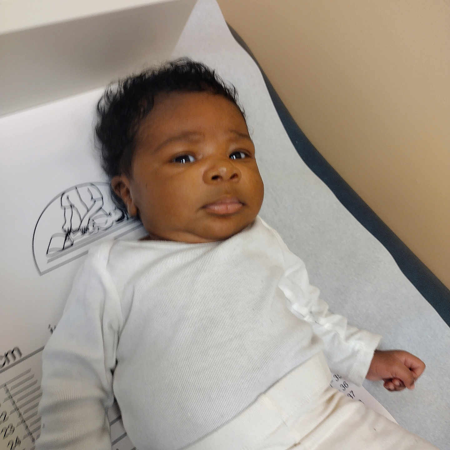 Noah is registered to the contest to win money with this photo: baby, infant, child, medical, examination_table, white_clothing, lying_down, curious_expression, indoors, healthcare, clinic, person, skin, head, face, looking, young, pediatric, patient, casual