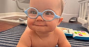 Jonah joined the competition — help win amazing prizes! baby, child, glasses, blue_glasses, smile, grinning, striped_rug, nursery, toy, bracelet, floor, indoor, infant, crawling, cute, playtime, furniture, window_blinds, soft_lighting, happy